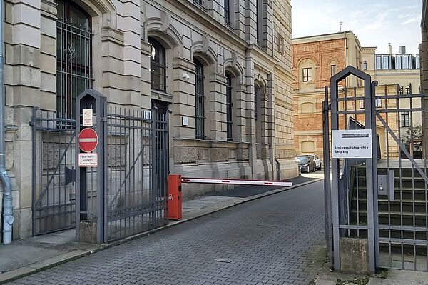 Das Foto zeigt ein historisches Gebäude mit einer Fassade aus hellem Stein und stilisierten Bögen über den Fenstern. Links ist ein schmiedeeisernes Tor mit einem Hinweisschild zu sehen, das den Zugang als Feuerwehreinfahrt kennzeichnet. Eine Schranke blockiert die Einfahrt, und auf der rechten Seite befindet sich ein Schild, das das Gebäude als Archiv der Universität Leipzig kennzeichnet.