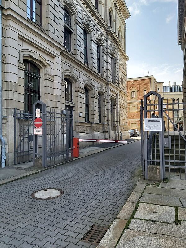 Das Foto zeigt ein historisches Gebäude mit einer Fassade aus hellem Stein und stilisierten Bögen über den Fenstern. Links ist ein schmiedeeisernes Tor mit einem Hinweisschild zu sehen, das den Zugang als Feuerwehreinfahrt kennzeichnet. Eine Schranke blockiert die Einfahrt, und auf der rechten Seite befindet sich ein Schild, das das Gebäude als Archiv der Universität Leipzig kennzeichnet.