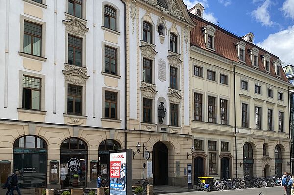 Das Bild zeigt eine Straßenszene mit mehreren historischen Gebäuden, die reich an architektonischen Verzierungen sind. Die Fassaden sind eine Mischung aus Weiß und Beige mit großen Fenstern und dekorativen Stuckarbeiten. Ein Gebäude hat eine Statue in einer Nische über dem Eingang. Entlang des Bürgersteigs sind Fahrräder geparkt, und es sind einige Menschen zu Fuß unterwegs. Im Vordergrund befindet sich ein digitales Werbeschild.