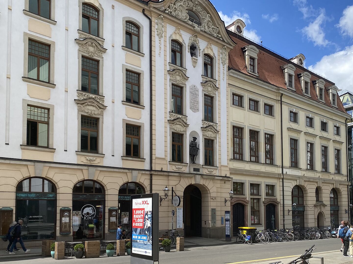 Das Bild zeigt eine Straßenszene mit mehreren historischen Gebäuden, die reich an architektonischen Verzierungen sind. Die Fassaden sind eine Mischung aus Weiß und Beige mit großen Fenstern und dekorativen Stuckarbeiten. Ein Gebäude hat eine Statue in einer Nische über dem Eingang. Entlang des Bürgersteigs sind Fahrräder geparkt, und es sind einige Menschen zu Fuß unterwegs. Im Vordergrund befindet sich ein digitales Werbeschild.