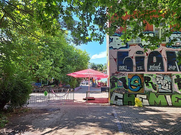Das Bild zeigt den Eingangsbereich von Conne Island, einem Kulturzentrum in Leipzig. Das Gebäude ist mit bunten Graffitis bedeckt. Im Vordergrund stehen Tische und rote Sonnenschirme, die von Bäumen umgeben sind. Ein Metallzaun mit dekorativen Bändern markiert den Eingang. Im Hintergrund sind weitere Graffitis und Bäume zu sehen.