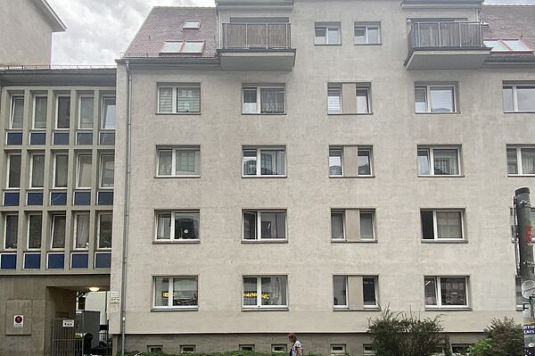 Sie Hochgeladenes Bild Copilot Das Bild zeigt die Fassade eines mehrstöckigen Wohngebäudes. Das Gebäude hat mehrere Fenster, die in einem Gittermuster über die Fassade verteilt sind. Im obersten Stockwerk befinden sich zwei Balkone und auf dem Dach gibt es Dachfenster. Vor dem Gebäude sind einige Fahrräder abgestellt. Am Fuß des Gebäudes gibt es eine kleine Gartenfläche mit Büschen und Grünflächen. Auf der linken Seite des Bildes führt ein Durchgang zum Inneren des Gebäudes.