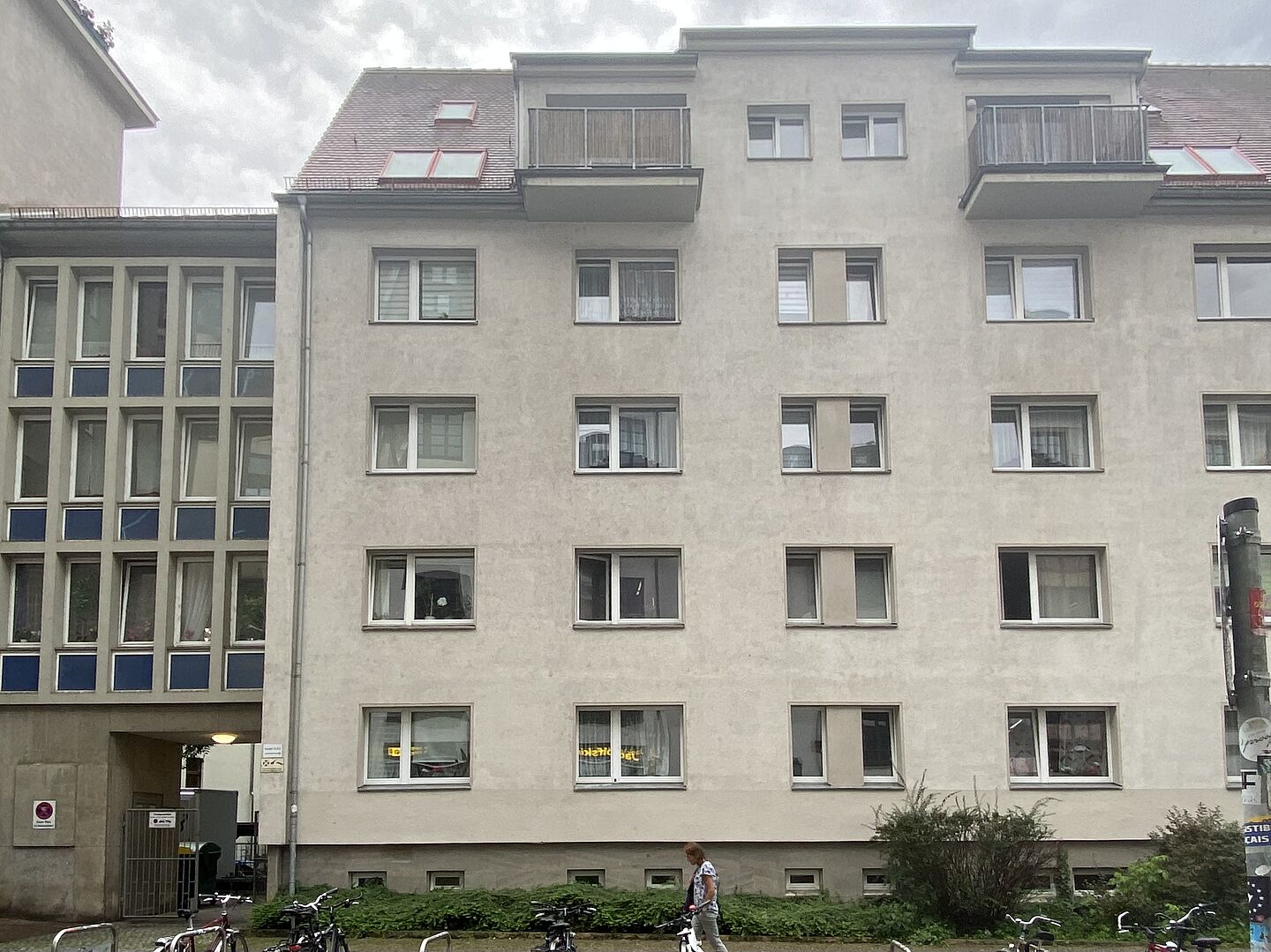 Sie Hochgeladenes Bild Copilot Das Bild zeigt die Fassade eines mehrstöckigen Wohngebäudes. Das Gebäude hat mehrere Fenster, die in einem Gittermuster über die Fassade verteilt sind. Im obersten Stockwerk befinden sich zwei Balkone und auf dem Dach gibt es Dachfenster. Vor dem Gebäude sind einige Fahrräder abgestellt. Am Fuß des Gebäudes gibt es eine kleine Gartenfläche mit Büschen und Grünflächen. Auf der linken Seite des Bildes führt ein Durchgang zum Inneren des Gebäudes.