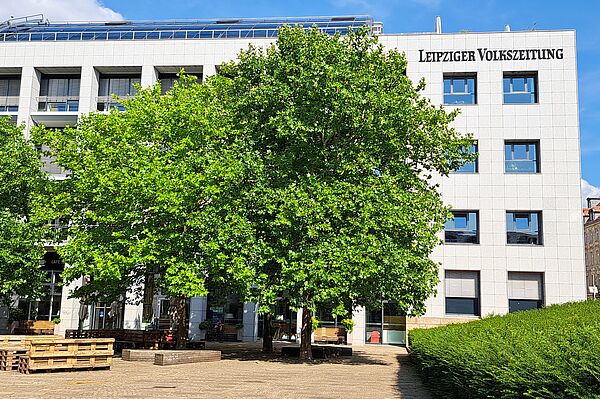 Das Bild zeigt das Gebäude der "Leipziger Volkszeitung", ein modernes Bauwerk mit einer Fassade aus hellen Steinplatten und rechteckigen Fenstern. Der Name der Zeitung ist deutlich an der oberen Fassade in großen Buchstaben angezeigt. Vor dem Gebäude befindet sich ein kleiner Platz mit gepflastertem Boden und einer Café-Terrasse, die von grünen Bäumen umgeben ist.