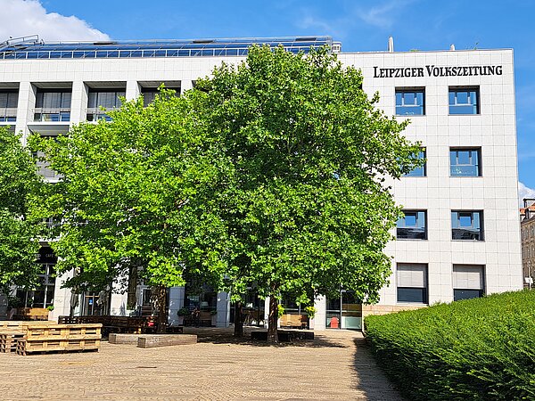 Das Bild zeigt das Gebäude der "Leipziger Volkszeitung", ein modernes Bauwerk mit einer Fassade aus hellen Steinplatten und rechteckigen Fenstern. Der Name der Zeitung ist deutlich an der oberen Fassade in großen Buchstaben angezeigt. Vor dem Gebäude befindet sich ein kleiner Platz mit gepflastertem Boden und einer Café-Terrasse, die von grünen Bäumen umgeben ist.