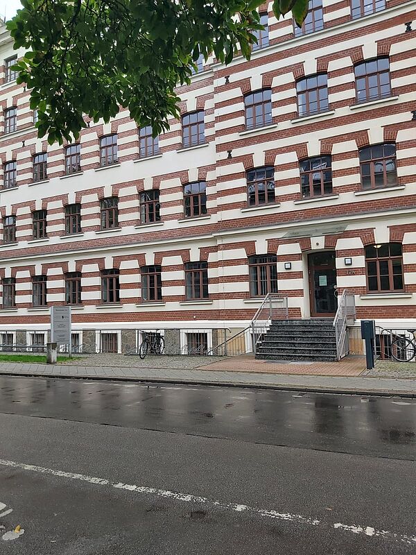 Das Bild zeigt die Regionalstelle Leipzig des Landesamtes für Schule und Bildung. Das Gebäude hat eine auffällige Fassade mit rot-weißen Ziegelmustern und großen Fenstern, die symmetrisch angeordnet sind. Der Eingangsbereich ist durch eine Treppe erreichbar, die zu einer Tür mit einem kleinen Vordach führt. Rechts und links vom Eingang stehen Fahrradständer.