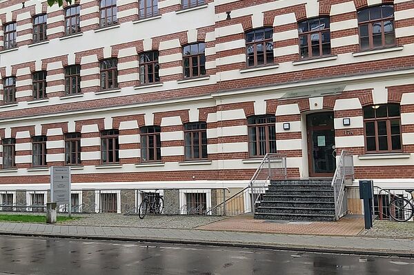 Das Bild zeigt die Regionalstelle Leipzig des Landesamtes für Schule und Bildung. Das Gebäude hat eine auffällige Fassade mit rot-weißen Ziegelmustern und großen Fenstern, die symmetrisch angeordnet sind. Der Eingangsbereich ist durch eine Treppe erreichbar, die zu einer Tür mit einem kleinen Vordach führt. Rechts und links vom Eingang stehen Fahrradständer.