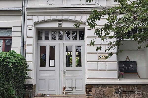 Das Bild zeigt den Eingang eines historischen Gebäudes mit einer weißen Fassade. Die Haustür ist zweiflügelig, aus Holz und in einem leicht abgenutzten Zustand. Oberhalb der Tür ist die Hausnummer „32“ angebracht. Das Gebäude weist klassische architektonische Elemente auf, wie Fensterumrandungen und ein dekoratives Gesims, was auf ein älteres Baujahr hinweist. Auf der rechten Seite befindet sich ein Schaufenster mit einer Tafel, auf der Zeichnungen oder Kunstwerke zu sehen sind. Eine niedrige Hecke wächst entlang der linken Seite des Gehwegs, der zum Eingang führt.