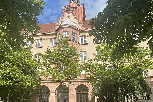 Das Bild zeigt ein großes, mehrstöckiges Gebäude mit beiger Fassade und roten Backsteinakzenten. Es hat ein rotes Ziegeldach, und im zentralen Bereich befindet sich ein kleiner Turm. Die Fenster sind symmetrisch angeordnet, und der Eingang ist von großen Bögen umrahmt. Um das Gebäude herum wachsen grüne Bäume, die den unteren Teil teilweise verdecken.