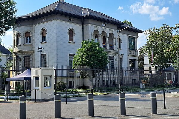 Das Bild zeigt das Generalkonsulat der Vereinigten Staaten von Amerika, ein markantes Gebäude mit klassischen architektonischen Merkmalen. Die Struktur weist eine helle Fassade mit Steinverzierungen auf, abgerundet durch ein Schieferdach. Architektonische Details wie Rundbogenfenster und ein Balkon mit dekorativem Geländer sind klar erkennbar. Ein amerikanischer Flaggenmast steht prominent vor dem Gebäude. Die Umgebung wird durch schützende Zäune und eine Wachhütte gesichert.