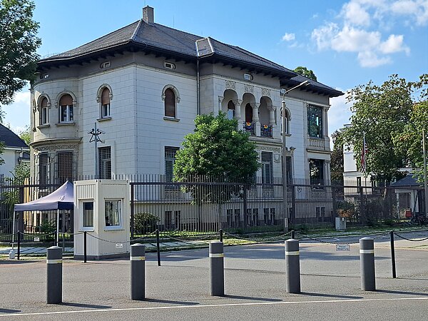 Das Bild zeigt das Generalkonsulat der Vereinigten Staaten von Amerika, ein markantes Gebäude mit klassischen architektonischen Merkmalen. Die Struktur weist eine helle Fassade mit Steinverzierungen auf, abgerundet durch ein Schieferdach. Architektonische Details wie Rundbogenfenster und ein Balkon mit dekorativem Geländer sind klar erkennbar. Ein amerikanischer Flaggenmast steht prominent vor dem Gebäude. Die Umgebung wird durch schützende Zäune und eine Wachhütte gesichert.