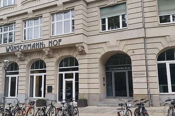 Das Bild zeigt ein Gebäude mit einer klassischen, sandfarbenen Fassade und eleganten Details. Es ist an einer Kopfsteinpflasterstraße gelegen und trägt den Schriftzug „WÜNSCHMANNS HOF“ prominent über dem Eingang. Im Erdgeschoss befinden sich Eingänge zu verschiedenen weiteren Einrichtungen.