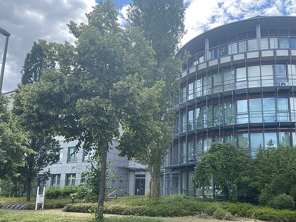 Das Bild zeigt ein modernes Gebäude mit einer geschwungenen Fassade und großen Glasfenstern. Mehrere Balkone sind ebenfalls zu sehen. Das Gebäude ist von üppigem Grün, einschließlich großer Bäume und Büsche, umgeben. Ein Weg führt zum Eingang des Gebäudes, und auf der linken Seite des Bildes ist ein Fahrradständer zu sehen.