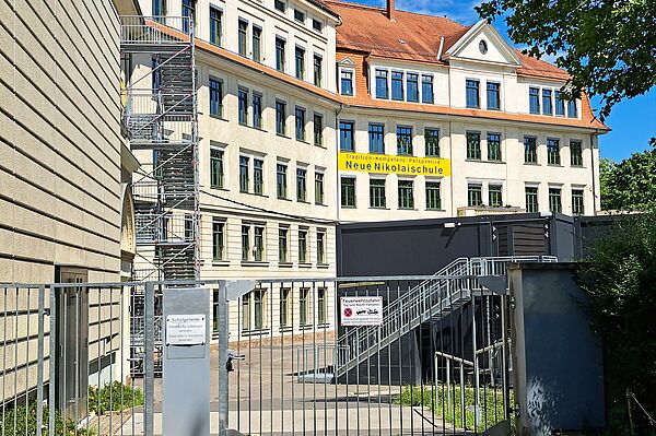 Das Bild zeigt ein großes, vierstöckiges Schulgebäude der "Neue Nikolaischule" mit traditioneller Architektur, einschließlich einem Giebeldach und symmetrisch angeordneten Fenstern. Das Gebäude ist in hellen Farbtönen gehalten. Im Vordergrund ist ein graues Metalltor zu sehen, das Zugang zum Schulgelände gewährt. Rechts neben dem Tor befindet sich eine Außentreppe, die zu einem Conatinergebäude führt. Der Himmel ist blau und der Vordergrund zeigt einige grüne Bäume, die teilweise das Gebäude verdecken.