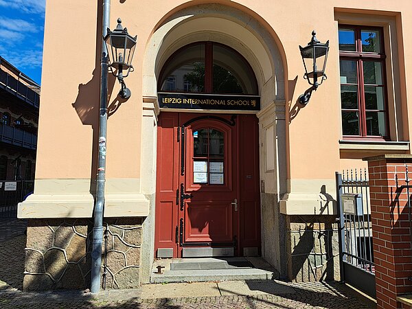 Das Bild zeigt den Eingangsbereich der Internationalen Schule Leipzig, gekennzeichnet durch eine große, bogige Eingangstür in Rot. Über dem Eingang befindet sich ein Schild mit der Aufschrift "LEIPZIG INTERNATIONAL SCHOOL". Das Gebäude selbst ist mit einer hellen Fassade versehen, ergänzt durch dekorative Steinelemente an der Basis. Zu beiden Seiten des Eingangs hängen klassische Laternen.