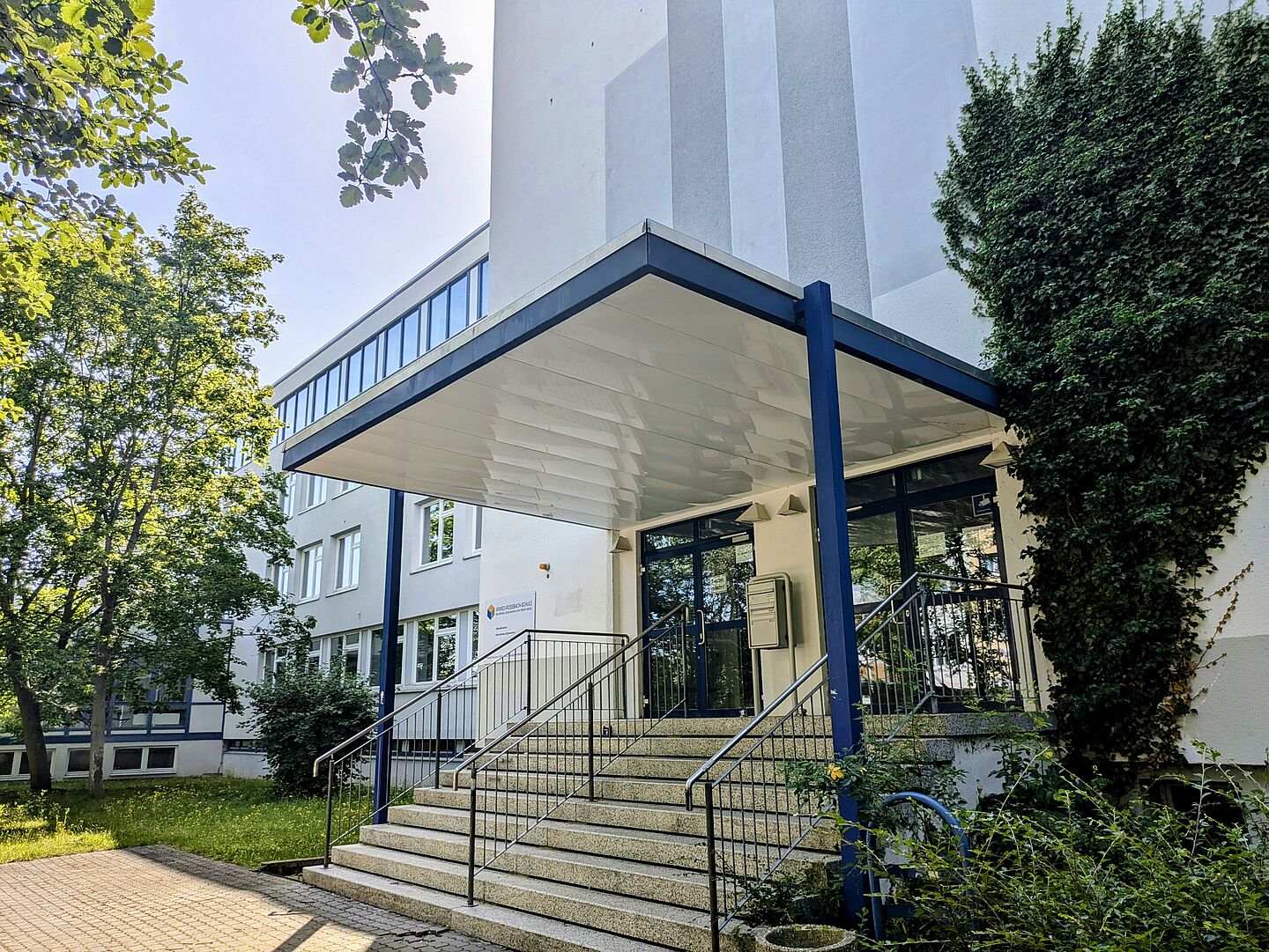 Das Bild zeigt die Arwed-Rossbach-Schule, ein modernes Gebäude mit einer weißen Fassade und großen Fenstern. Der Eingangsbereich hat ein Vordach, das von blauen Stützpfeilern getragen wird. Eine Treppe mit Geländern führt zum Eingang. Auf der rechten Seite ist die Fassade teilweise mit grünen Ranken bewachsen. Im Vordergrund und links sind Bäume und Grünflächen zu sehen.