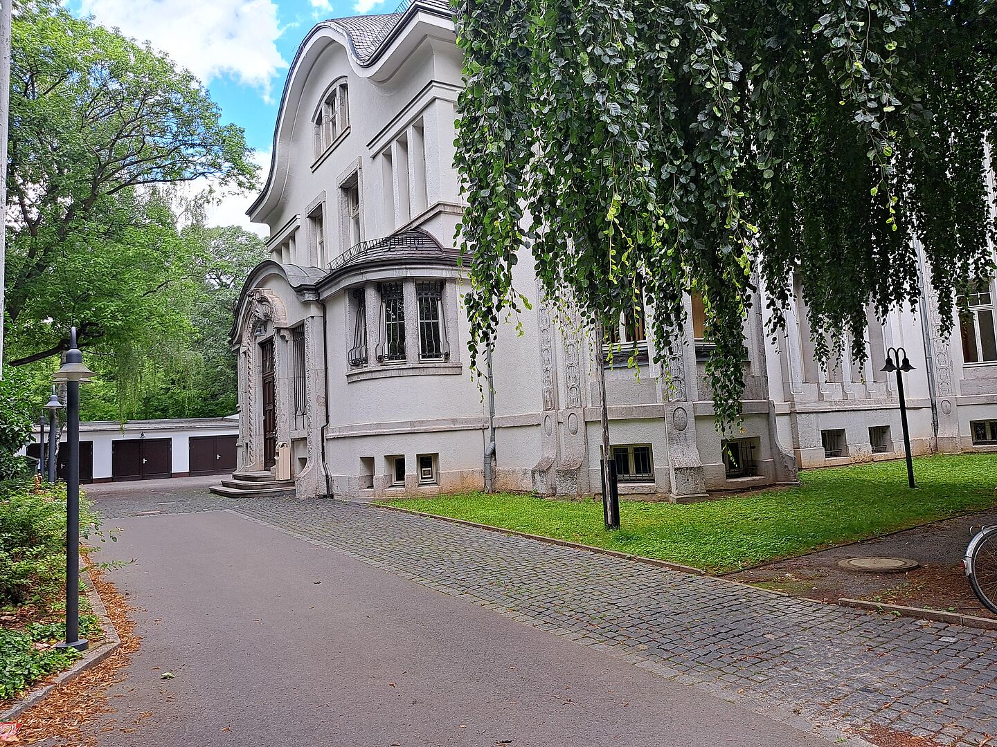 Das Bild zeigt das Deutsche Literaturinstitut, ein imposantes Gebäude mit einer weißen Fassade, umgeben von reichlich Grünfläche und Bäumen. Die Architektur des Gebäudes ist elegant mit geschwungenen Fenstern und einer dekorativen Eingangspartie, die durch eine überdachte Säulenstruktur gekennzeichnet ist. Die Fenster sind von künstlerischen Verzierungen umrahmt.