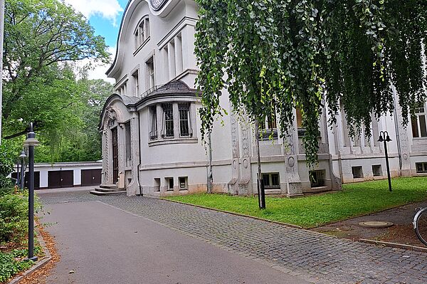 Das Bild zeigt das Deutsche Literaturinstitut, ein imposantes Gebäude mit einer weißen Fassade, umgeben von reichlich Grünfläche und Bäumen. Die Architektur des Gebäudes ist elegant mit geschwungenen Fenstern und einer dekorativen Eingangspartie, die durch eine überdachte Säulenstruktur gekennzeichnet ist. Die Fenster sind von künstlerischen Verzierungen umrahmt.