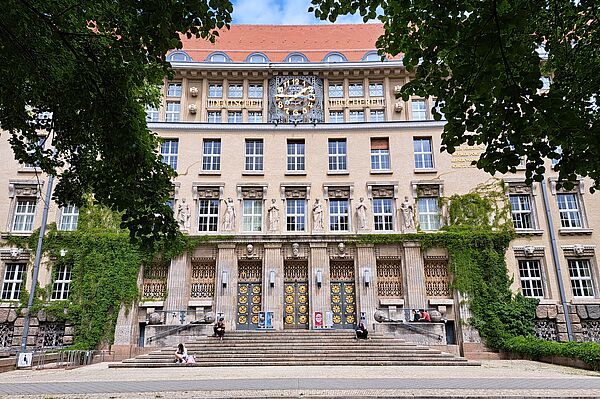 Das Bild zeigt den Haupteingang der Deutschen Nationalbibliothek in Leipzig. Das historische Gebäude hat große Fenster, Statuen und eine Uhr über dem Eingang mit dem Schriftzug "Deutsche National Bibliothek". Die Fassade ist teils mit Efeu bewachsen. Breite Treppen führen zu verzierten Türen, vor denen einige Menschen sitzen. Der Vorplatz ist gepflastert und von Bäumen umrahmt.