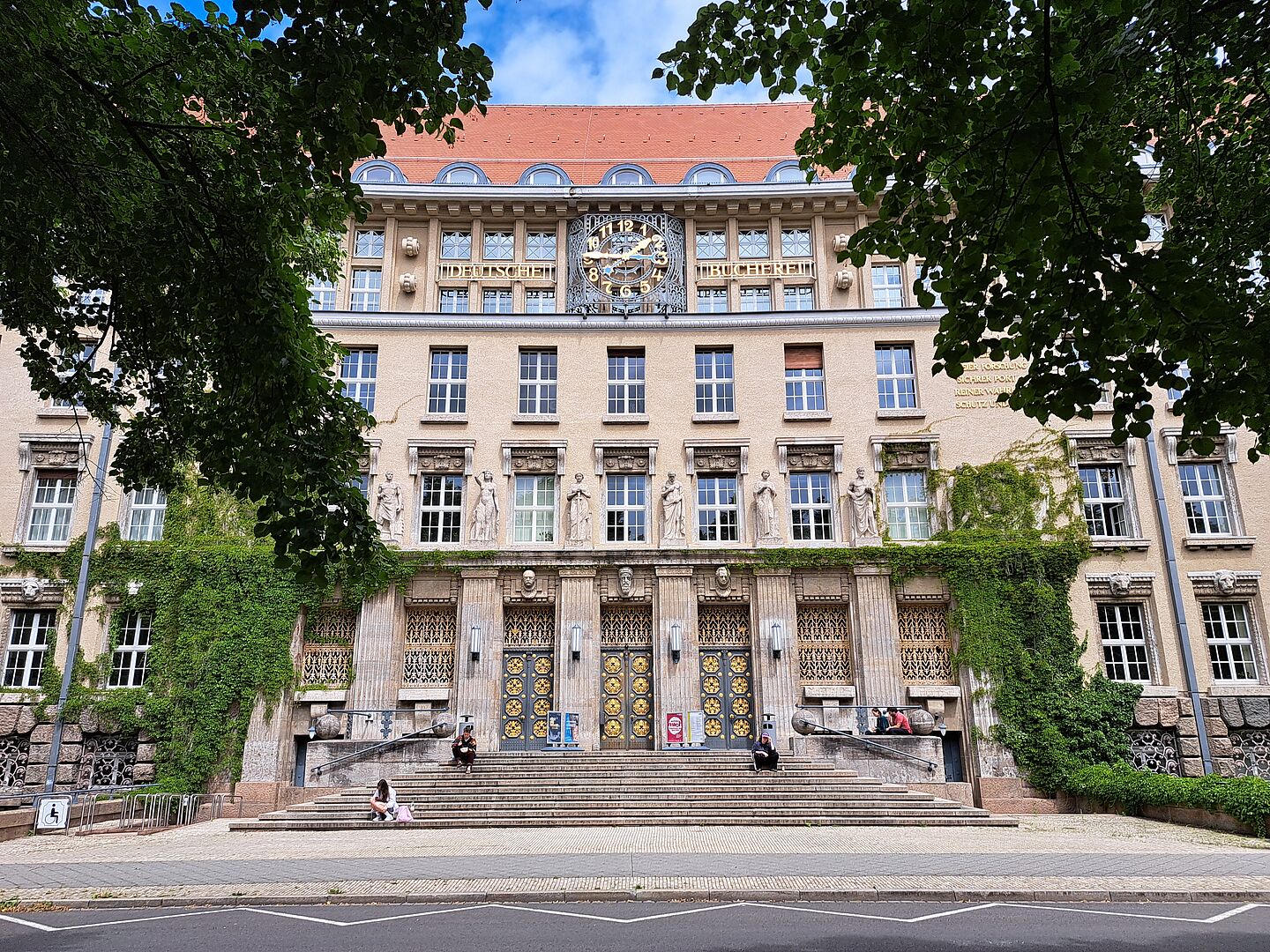 Das Bild zeigt den Haupteingang der Deutschen Nationalbibliothek in Leipzig. Das historische Gebäude hat große Fenster, Statuen und eine Uhr über dem Eingang mit dem Schriftzug "Deutsche National Bibliothek". Die Fassade ist teils mit Efeu bewachsen. Breite Treppen führen zu verzierten Türen, vor denen einige Menschen sitzen. Der Vorplatz ist gepflastert und von Bäumen umrahmt.