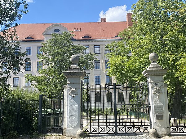  Das Bild zeigt den Eingang eines großen Gebäudes mit einem roten Ziegeldach und mehreren Fenstern. Der Eingang wird von zwei steinernen Säulen eingerahmt, die oben mit kugelförmigen Ornamenten verziert sind. Die Säulen sind Teil eines eisernen Tores. Um den Eingang herum gibt es Bäume und Grünflächen, die teilweise die Sicht auf das Gebäude verdecken. Die Fassade des Gebäudes ist symmetrisch, mit einem zentralen Abschnitt, der ein bogenförmiges Fenster oben beinhaltet. 