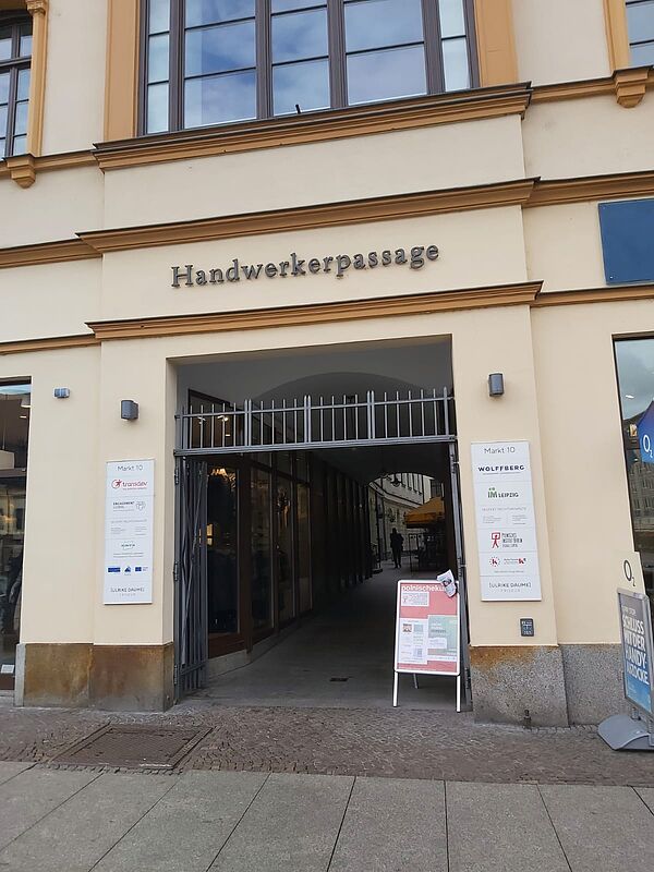 Das Bild zeigt den Eingang zur "Handwerkerpassage" in einem historischen Gebäude. Über dem Torbogen ist der Name "Handwerkerpassage" in großen, metallisch glänzenden Buchstaben angebracht. Es handelt sich um einen Durchgang, der von außen zugänglich ist, und hinter dem Eingang scheint eine schmale Passage mit Geschäften oder Büros zu verlaufen.  Links vom Eingang befindet sich ein Schild mit mehreren Logos und Namen, die vermutlich auf Unternehmen oder Institutionen hinweisen, die in diesem Gebäude ansässig sind.