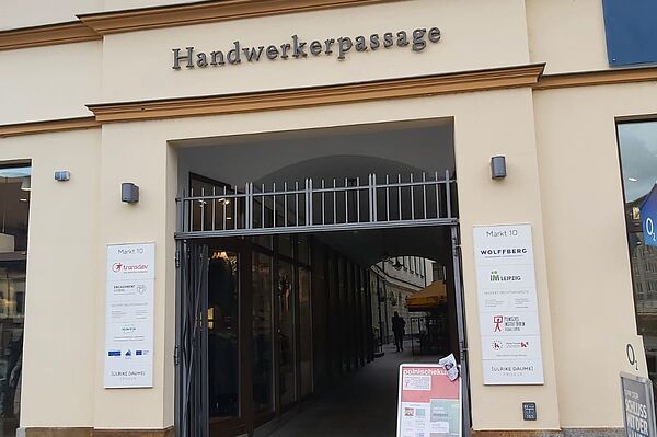 Das Bild zeigt den Eingang zur "Handwerkerpassage" in einem historischen Gebäude. Über dem Torbogen ist der Name "Handwerkerpassage" in großen, metallisch glänzenden Buchstaben angebracht. Es handelt sich um einen Durchgang, der von außen zugänglich ist, und hinter dem Eingang scheint eine schmale Passage mit Geschäften oder Büros zu verlaufen.  Links vom Eingang befindet sich ein Schild mit mehreren Logos und Namen, die vermutlich auf Unternehmen oder Institutionen hinweisen, die in diesem Gebäude ansässig sind.