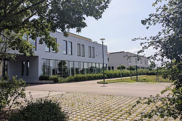 Die Außenansicht der Bibliothek der Berufsakademie Sachsen - Staatliche Studienakademie Leipzig zeigt ein modernes zweistöckiges Gebäude mit einer hellgrauen Fassade und großen Fensterfronten. Vor dem Gebäude befinden sich gepflasterte Parkplätze und eine Straßenlaterne. Ein großer Baum und einige Sträucher im Vordergrund sowie eine Grünflächen und kleine Bäume umgeben das Gebäude. 