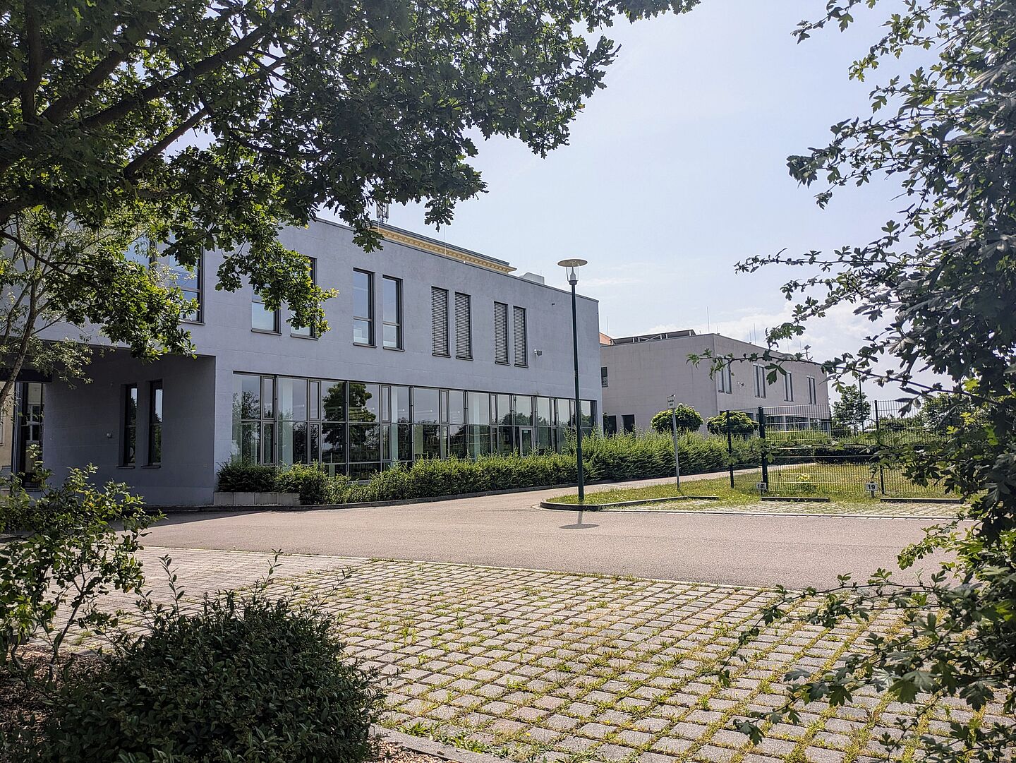 Die Außenansicht der Bibliothek der Berufsakademie Sachsen - Staatliche Studienakademie Leipzig zeigt ein modernes zweistöckiges Gebäude mit einer hellgrauen Fassade und großen Fensterfronten. Vor dem Gebäude befinden sich gepflasterte Parkplätze und eine Straßenlaterne. Ein großer Baum und einige Sträucher im Vordergrund sowie eine Grünflächen und kleine Bäume umgeben das Gebäude. 