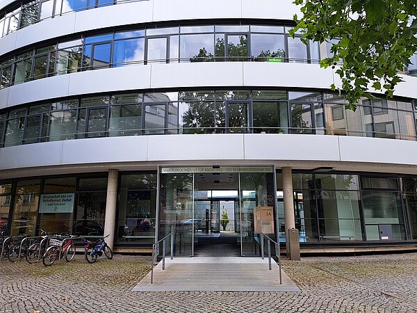 Das Bild zeigt das Max-Planck-Institut für Kognitions- und Neurowissenschaften, ein modernes Gebäude mit einer abgerundeten, mehrstöckigen Glasfassade. Die Struktur ist in helle, neutrale Farbtöne gehalten und bietet durch die großen Fenster viel natürliche Beleuchtung. Im Eingangsbereich befindet sich eine breite, zugängliche Tür, über der der Name des Instituts angezeigt wird. Vor dem Gebäude stehen mehrere Fahrräder, und eine Rampe führt direkt zum Haupteingang
