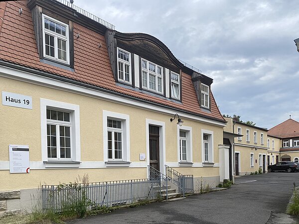 Sie Hochgeladenes Bild Copilot Das Bild zeigt ein Gebäude mit der Aufschrift "Haus 19." Die Fassade ist gelb, die Fensterrahmen weiß und das Dach mit roten Ziegeln gedeckt. Es gibt mehrere Fenster im Erdgeschoss und im oberen Stockwerk, welches Dachfenster besitzt. Eine kleine Treppe führt zu einer Tür im Erdgeschoss. An der Wand neben der Tür befindet sich ein Schild, ein weiteres Schild ist auf der linken Seite des Gebäudes sichtbar. Im Hintergrund ist ein weiteres ähnliches Gebäude zu sehen.