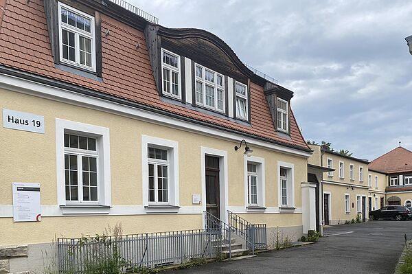 Sie Hochgeladenes Bild Copilot Das Bild zeigt ein Gebäude mit der Aufschrift "Haus 19." Die Fassade ist gelb, die Fensterrahmen weiß und das Dach mit roten Ziegeln gedeckt. Es gibt mehrere Fenster im Erdgeschoss und im oberen Stockwerk, welches Dachfenster besitzt. Eine kleine Treppe führt zu einer Tür im Erdgeschoss. An der Wand neben der Tür befindet sich ein Schild, ein weiteres Schild ist auf der linken Seite des Gebäudes sichtbar. Im Hintergrund ist ein weiteres ähnliches Gebäude zu sehen.