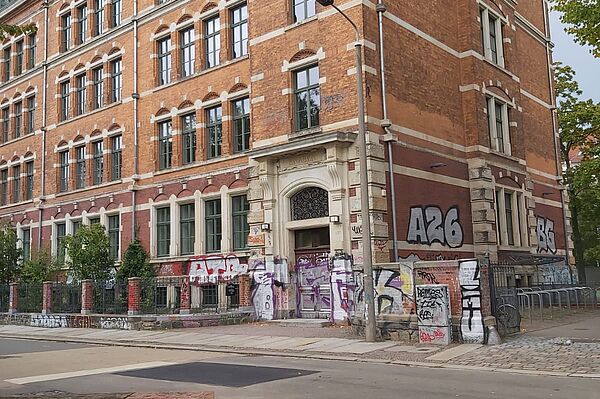 Das Bild zeigt die Fassade eines großen, historischen Backsteingebäudes. Es handelt sich um ein mehrstöckiges Gebäude aus roten Ziegelstein mit hellen Steinverzierungen. Besonders auffällig sind die zahlreichen Fenster, die dem Gebäude ein großzügiges und zugleich monumentales Erscheinungsbild verleihen. Der Eingangsbereich ist mit einem kunstvoll verzierten Bogen ausgestattet. Allerdings ist der untere Teil der Fassade, einschließlich des Eingangsbereichs, stark mit Graffiti bedeckt, was einen auffälligen Kontrast zu der sonst klassischen Architektur darstellt.