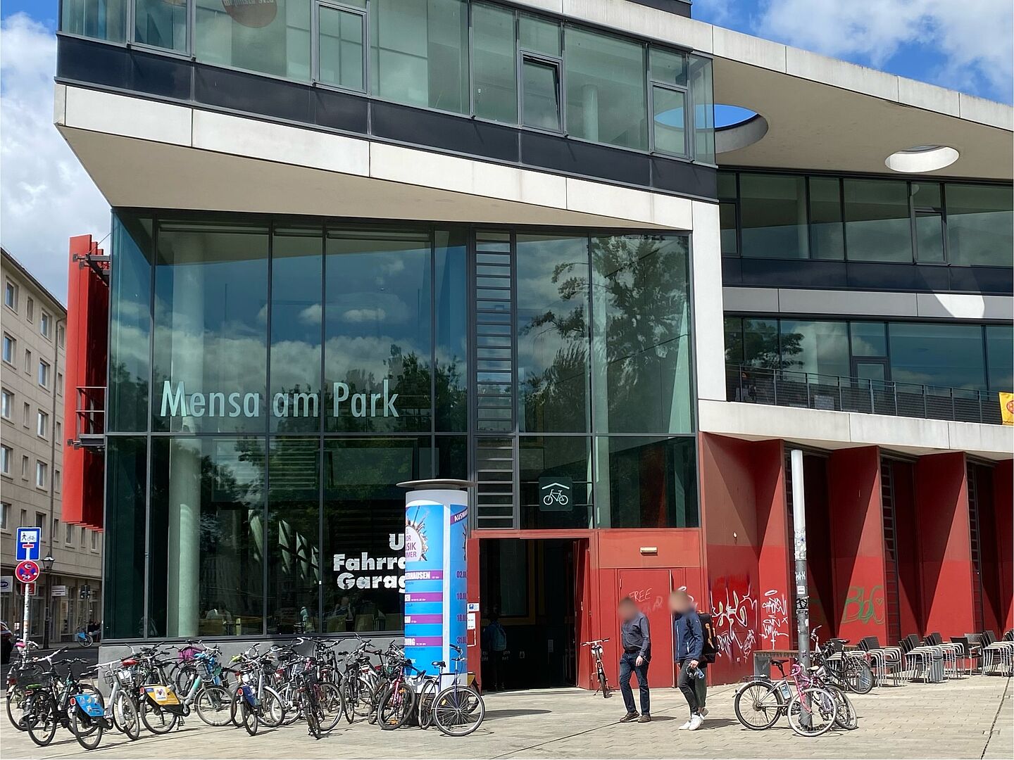 Ein modernes Gebäude mit großen Glasfronten und roten Akzentwänden. Über dem Eingang steht "Mensa am Park". Der große Eingang ist deutlich zu sehen und hat keine Türen. Dieser führt zur Fahrradgerage.