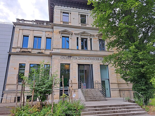 Das Bild zeigt die Galerie für Zeitgenössische Kunst, ein dreistöckiges Gebäude mit einer Fassade aus hellem Stein. Das Gebäude kombiniert klassische Architekturmerkmale wie Säulen und Reliefverzierungen mit modernen Elementen, erkennbar an den großflächigen Fenstern und dem schlichten, zeitgenössischen Schriftzug an der Fassade. Eine Treppe führt zum Haupteingang. Das Gebäude ist von grünen Bäumen umgeben.