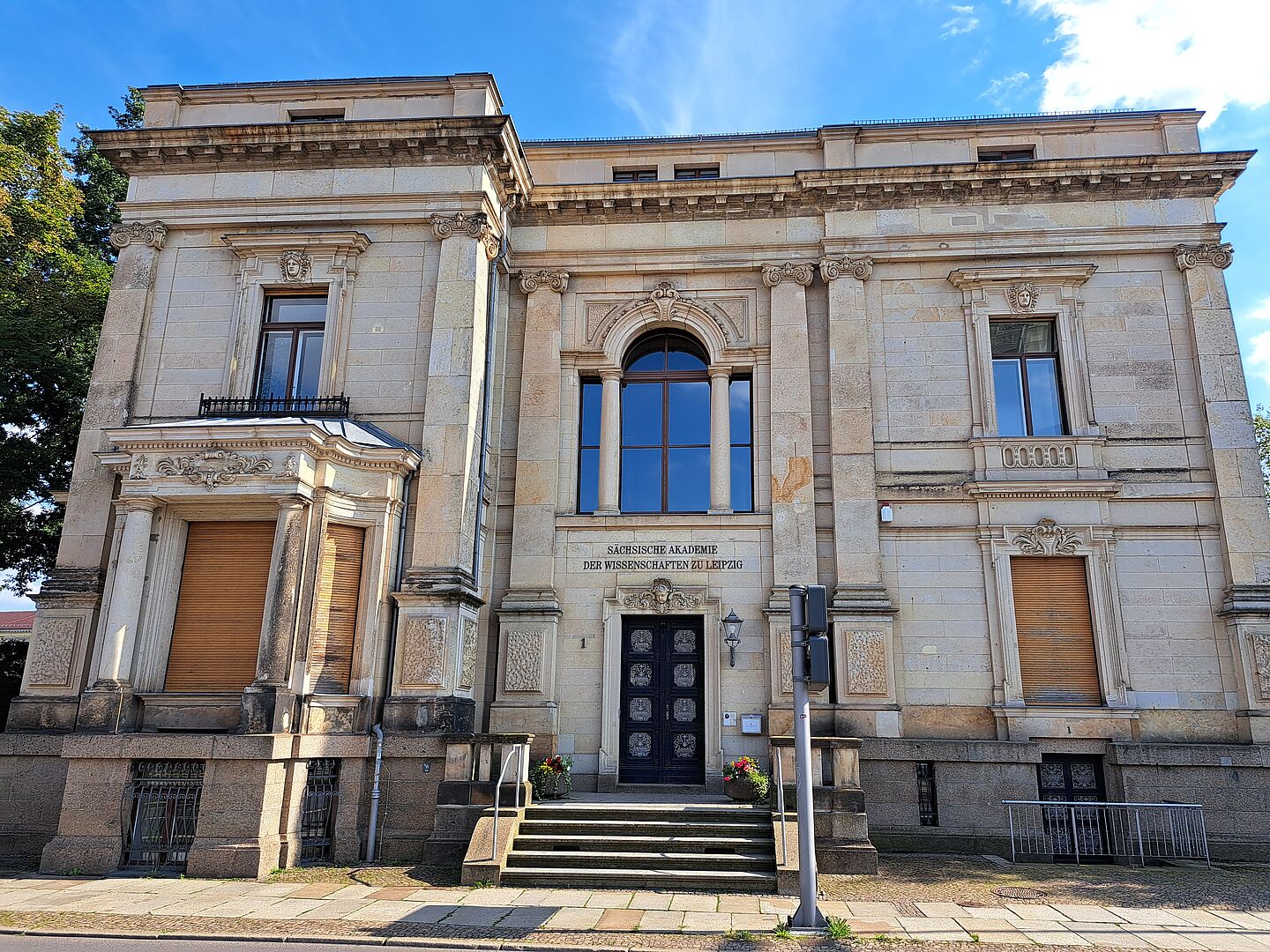 Das Bild zeigt die Sächsische Akademie der Wissenschaften zu Leipzig, ein beeindruckendes Gebäude mit einer klassischen Steinarchitektur. Das Gebäude verfügt über einen zentralen Eingang, der durch eine große Steintreppe und eine beeindruckende Tür hervorgehoben wird. Über der Tür befindet sich ein aufwendig gestaltetes Fenster, umrahmt von Steinsäulen und Skulpturen.