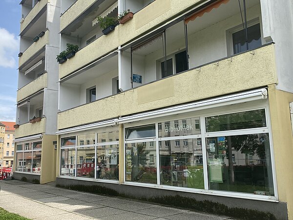 Leipziger Stadtteilbibliothek Schönefeld Das Bild zeigt die Fassade eines mehrstöckigen Gebäudes mit Balkonen. Im Erdgeschoss des Gebäudes befinden sich große Fenster, und ein Schild mit der Aufschrift "Bibliothek" weist darauf hin, dass sich dort eine Bibliothek befindet. Auf den Balkonen sind verschiedene Pflanzen und Blumentöpfe zu sehen. Das Gebäude liegt in einer städtischen Gegend, im Hintergrund sind weitere Gebäude zu erkennen.