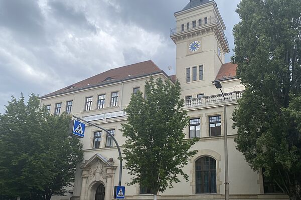 Das Bild zeigt die Friedrich-Schiller-Schule, ein eindrucksvolles historisches Gebäude mit einem markanten Turm, der eine große Uhr trägt. Das Gebäude hat eine cremefarbene Fassade und ein rotes Ziegeldach. Auf der Fassade ist ein Spruch zu sehen: "Schone fremde Freiheit, Zeige selbst Freiheit." Vor dem Gebäude stehen mehrere Bäume, die Teile der Fassade verdecken. Der Eingang des Gebäudes ist durch ein dekoratives Tor mit Steinornamenten hervorgehoben.