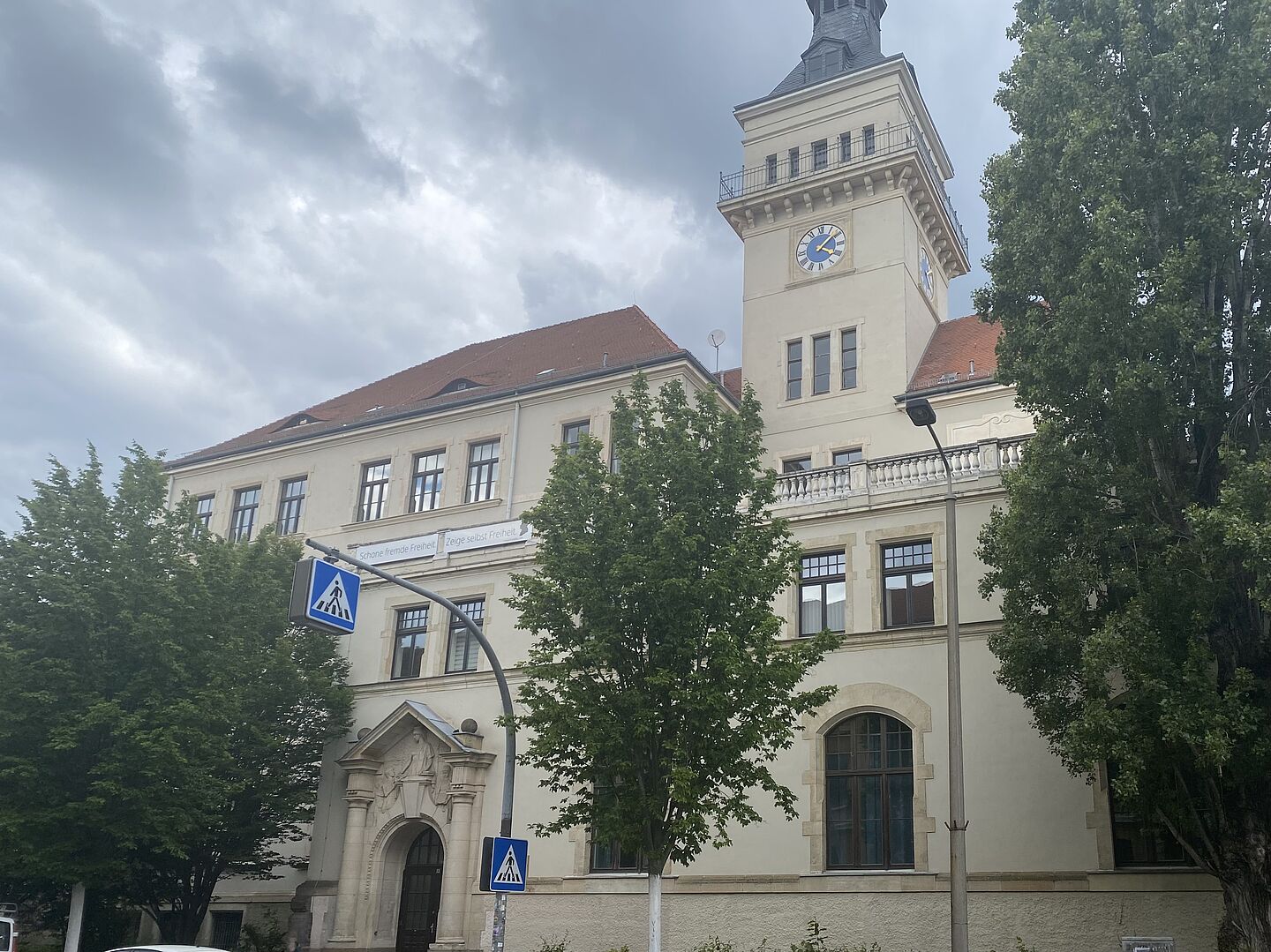 Das Bild zeigt die Friedrich-Schiller-Schule, ein eindrucksvolles historisches Gebäude mit einem markanten Turm, der eine große Uhr trägt. Das Gebäude hat eine cremefarbene Fassade und ein rotes Ziegeldach. Auf der Fassade ist ein Spruch zu sehen: "Schone fremde Freiheit, Zeige selbst Freiheit." Vor dem Gebäude stehen mehrere Bäume, die Teile der Fassade verdecken. Der Eingang des Gebäudes ist durch ein dekoratives Tor mit Steinornamenten hervorgehoben.